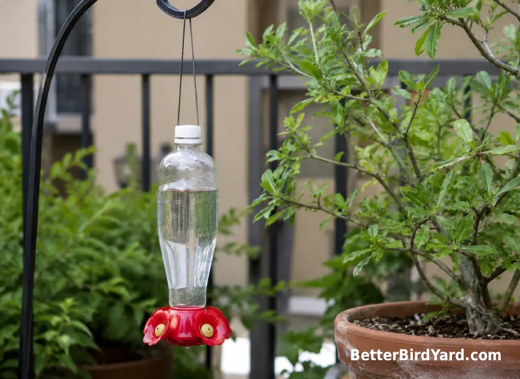 A small plastic bottle hummingbird feeder hangs from a shaded patio hook near potted greenery.
