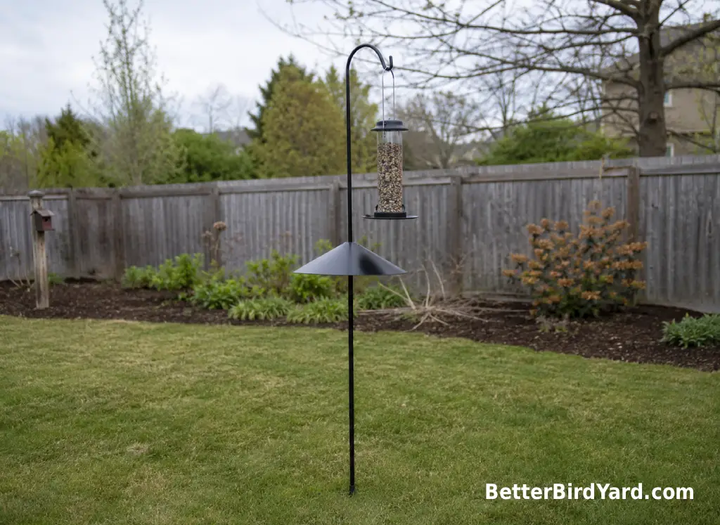 A bird feeder on a smooth pole stands away from a fence and shrubs in a suburban yard.