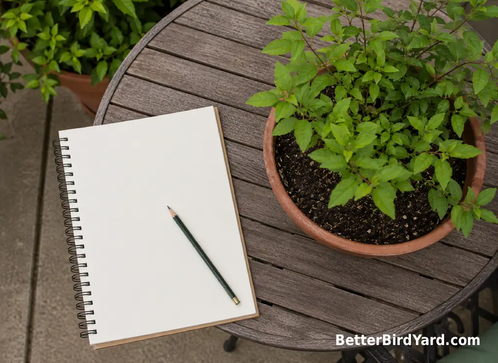 A sketchbook, pencil, and potted shrub sit on a patio table for backyard habitat planning.